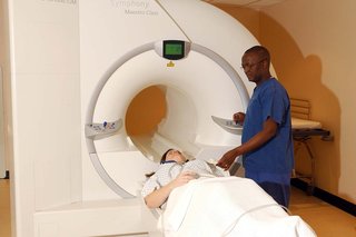 An MRI scanner with a short tunnel through the middle. A patient wearing a hospital gown lies on the narrow, flat bed that moves through the scanner. A healthcare professional is standing next to the patient.